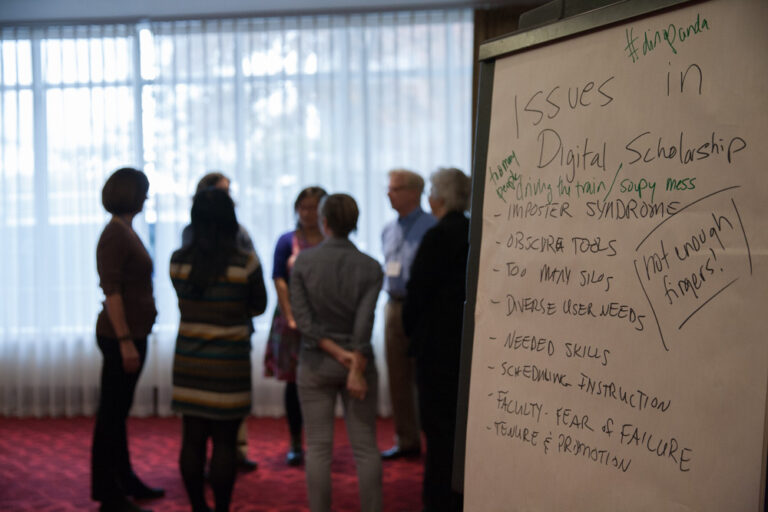 Foreground: poster pad listing "issues in digital scholarship" with attendees standing in a circle talking in the background